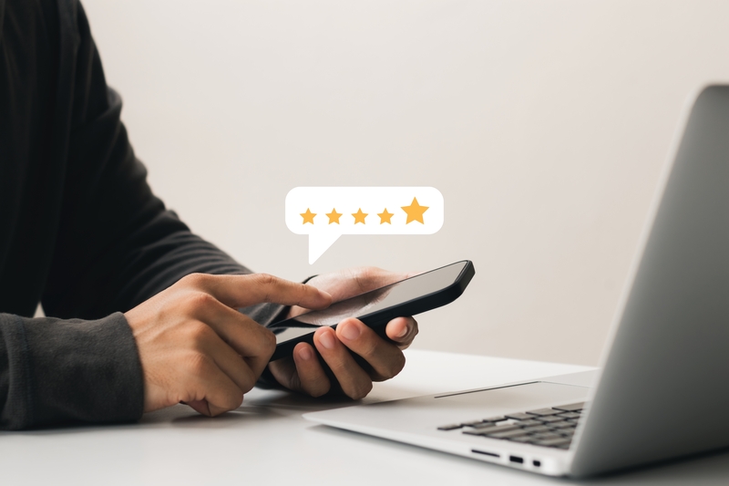 A male person’s hands, one holding a mobile phone in front of a Macbook, the other taps the phone’s screen to give a five-star review.