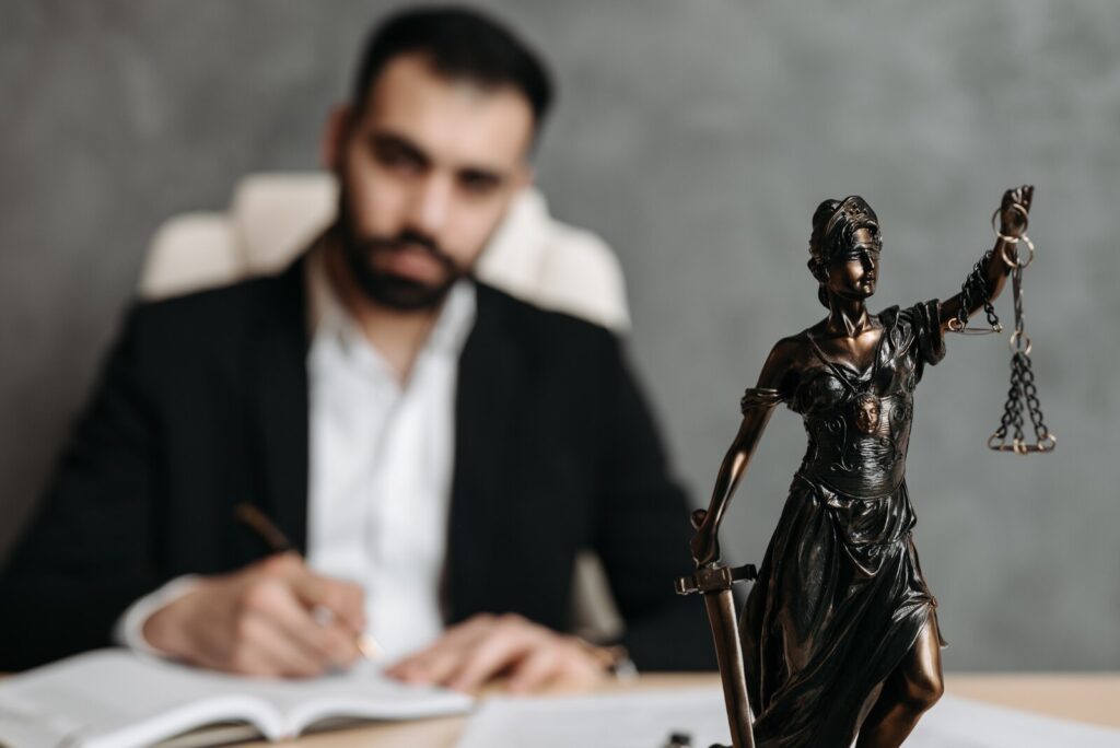 A copper lady justice figurine in focus. Out of focus in the background is a closed-bearded man wearing a white button-down and a black coat.